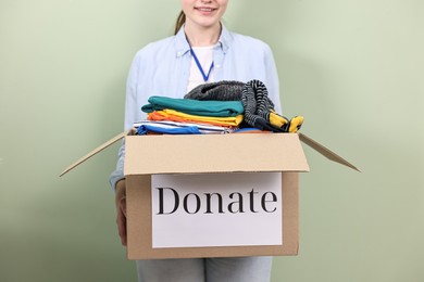 Photo of Woman holding donation box with clothes on pale olive background, closeup