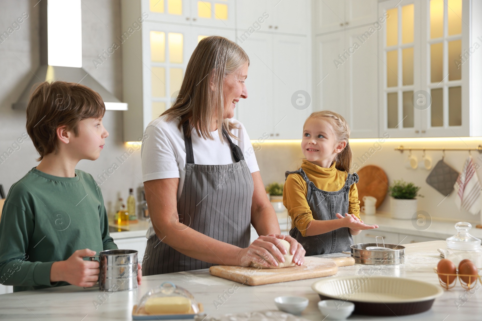 Photo of Grandmother and her grandchildren making dough at white marble table in kitchen