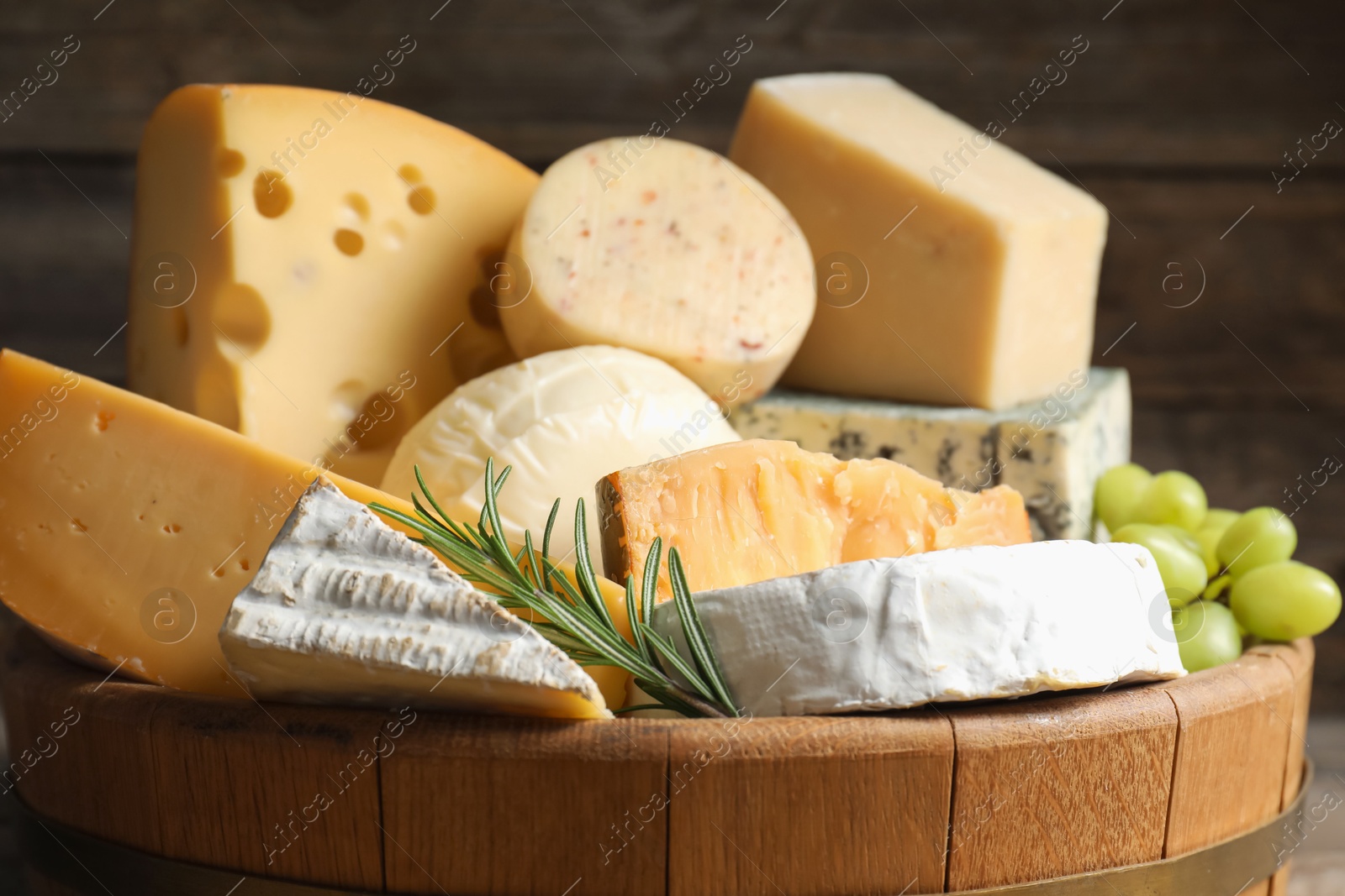 Photo of Different types of cheese, rosemary and grapes in wooden tray on blurred background, closeup