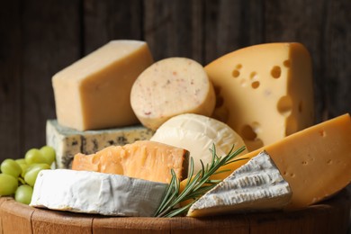 Photo of Different types of cheese, rosemary and grapes in wooden tray on blurred background, closeup