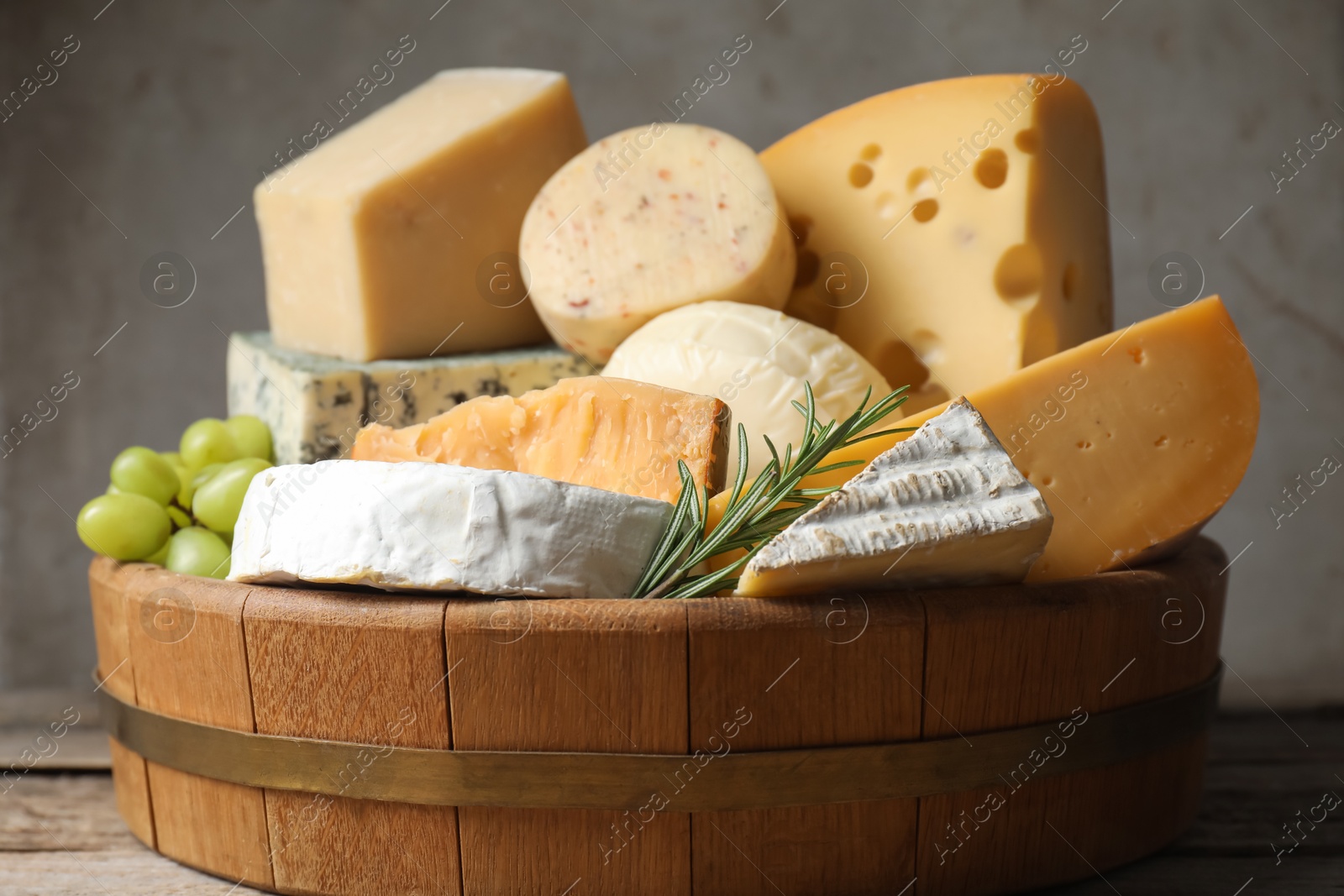 Photo of Different types of cheese, rosemary and grapes in wooden tray on table, closeup