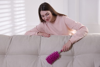 Photo of Happy woman cleaning sofa with duster at home