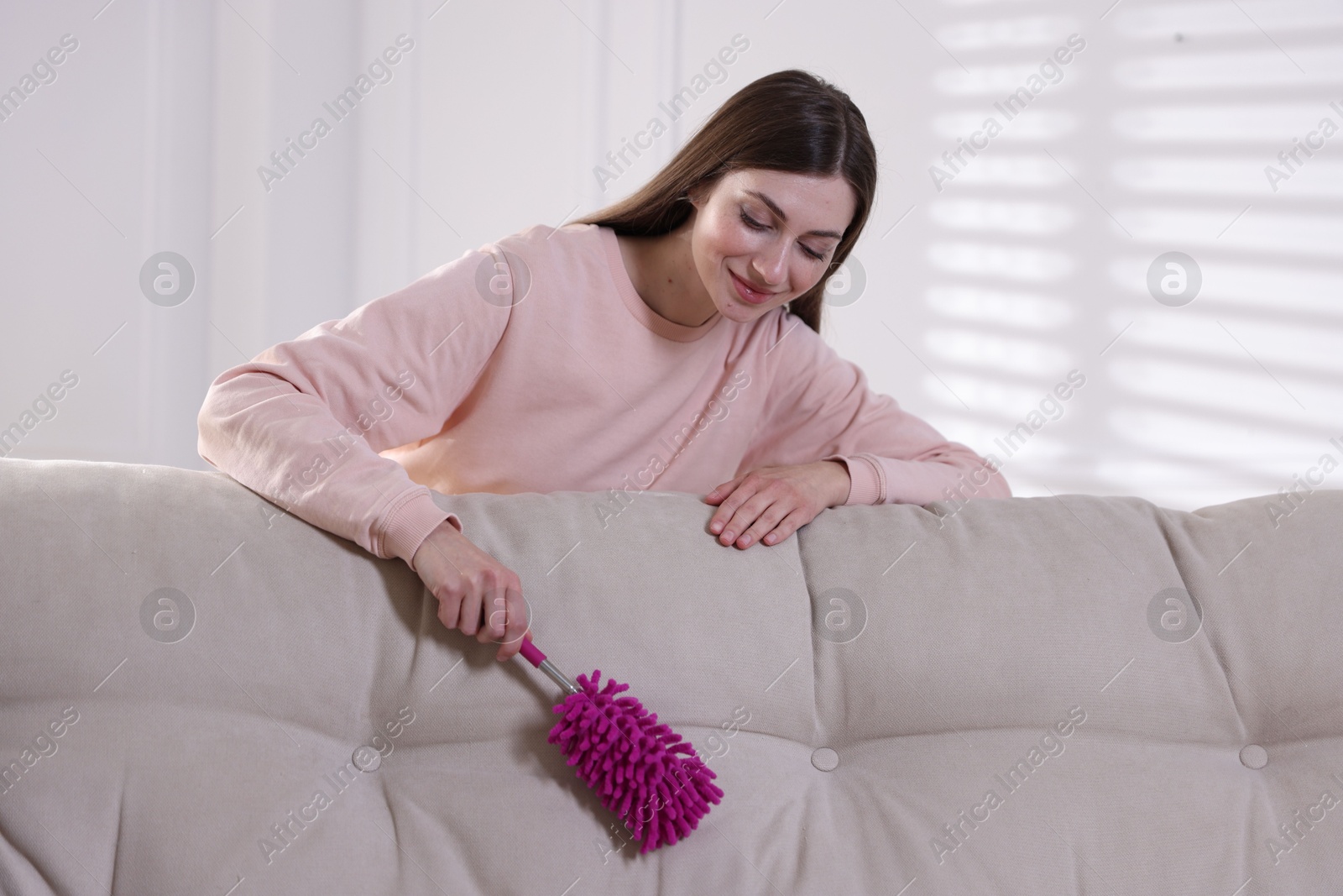 Photo of Happy woman cleaning sofa with duster at home