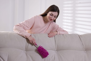 Photo of Happy woman cleaning sofa with duster at home