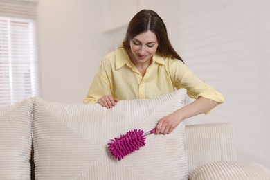 Photo of Happy woman cleaning sofa with duster at home