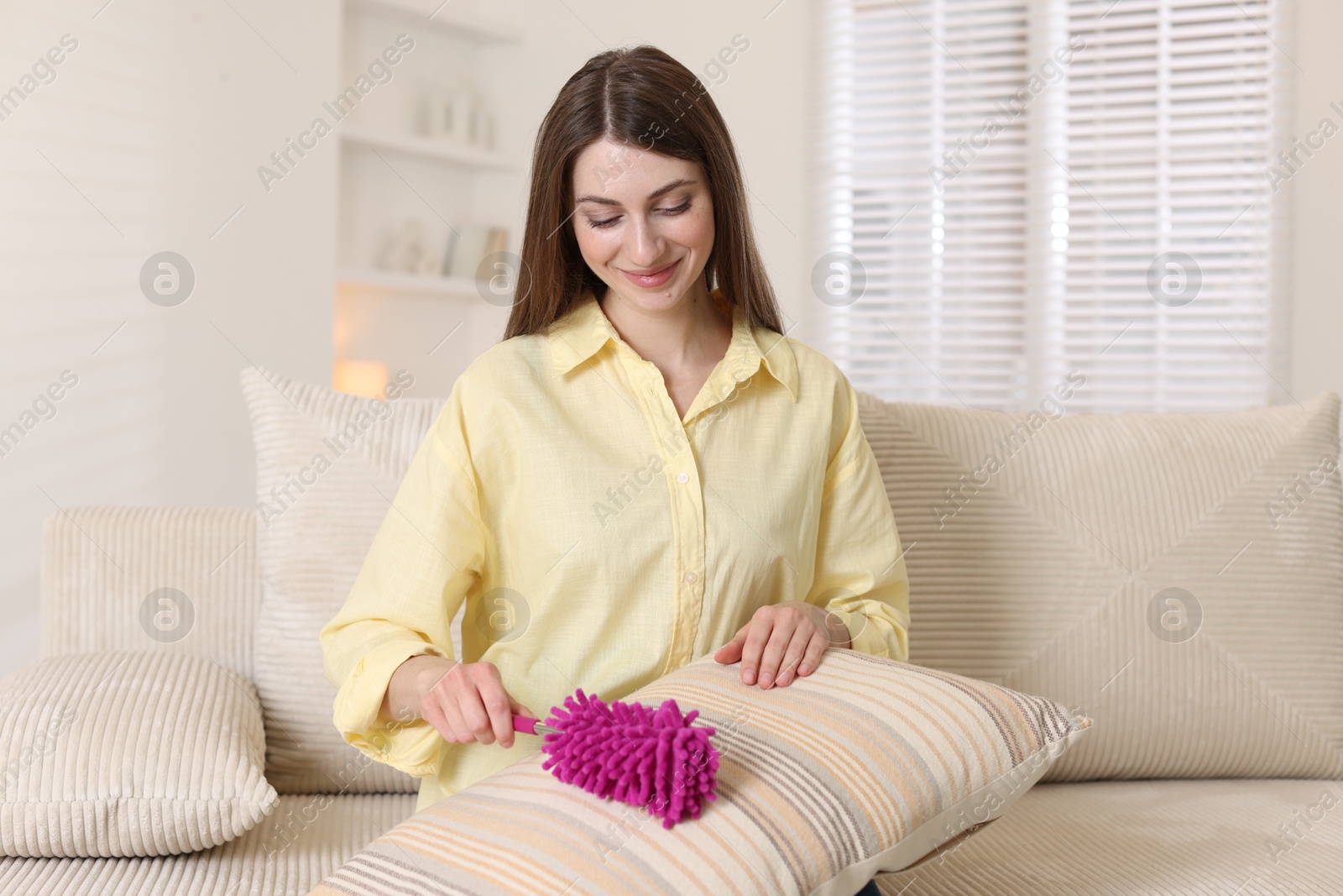 Photo of Happy woman cleaning sofa cushion with duster at home