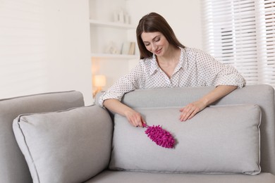 Photo of Happy woman cleaning sofa with duster at home