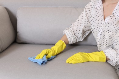 Photo of Woman in gloves cleaning sofa with rag at home, closeup