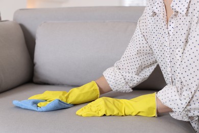 Photo of Woman in gloves cleaning sofa with rag at home, closeup