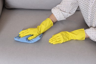 Photo of Woman in gloves cleaning sofa with rag at home, closeup