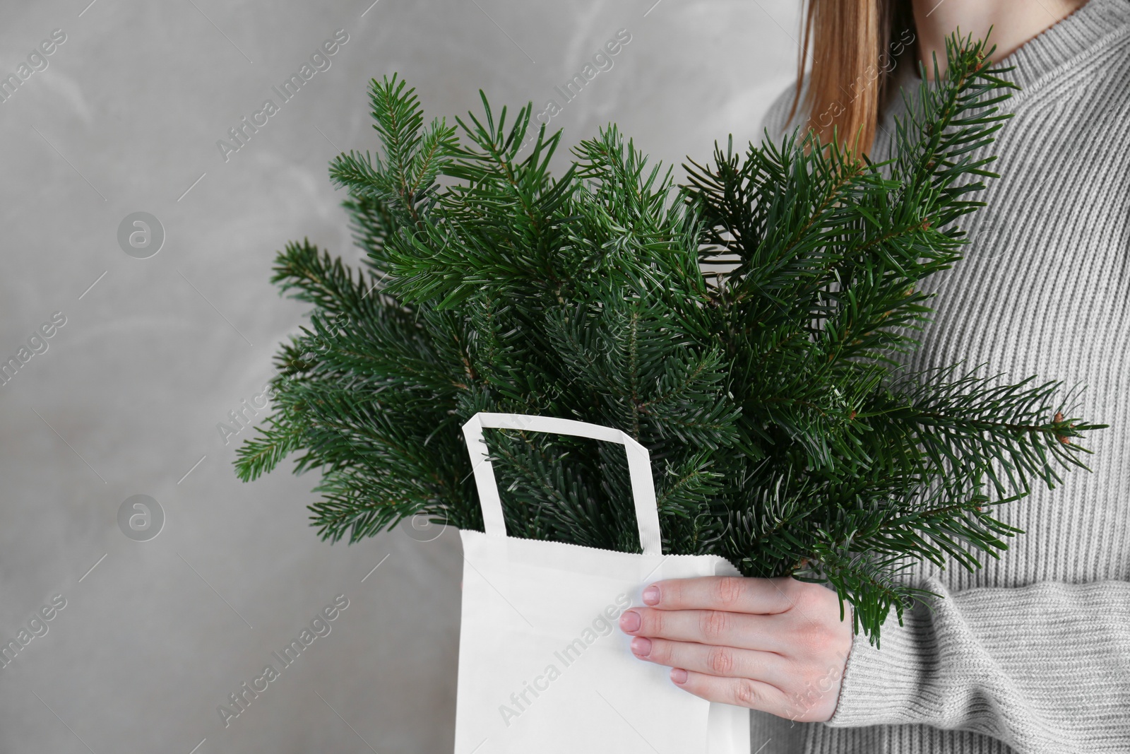 Photo of Woman holding green fir branches in paper bag near gray wall, closeup. Christmas decor