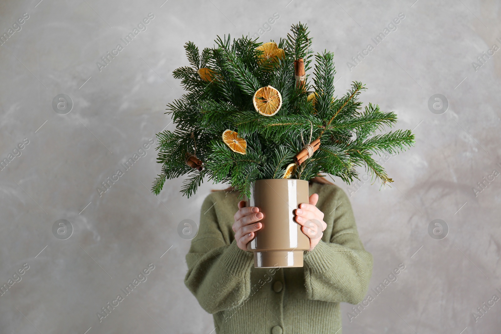 Photo of Woman holding beautiful Christmas composition of fir tree branches decorated with dried orange slices and cinnamon sticks near gray wall