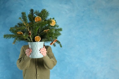 Photo of Woman holding beautiful Christmas composition of fir tree branches decorated with dried orange slices and cinnamon sticks near blue wall, space for text