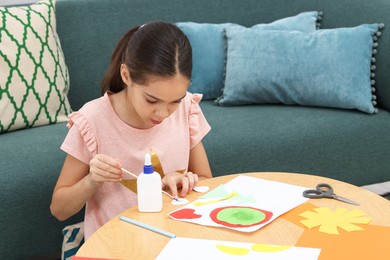 Photo of Girl applying glue onto paper figure for her creative project at table indoors. Art and craft