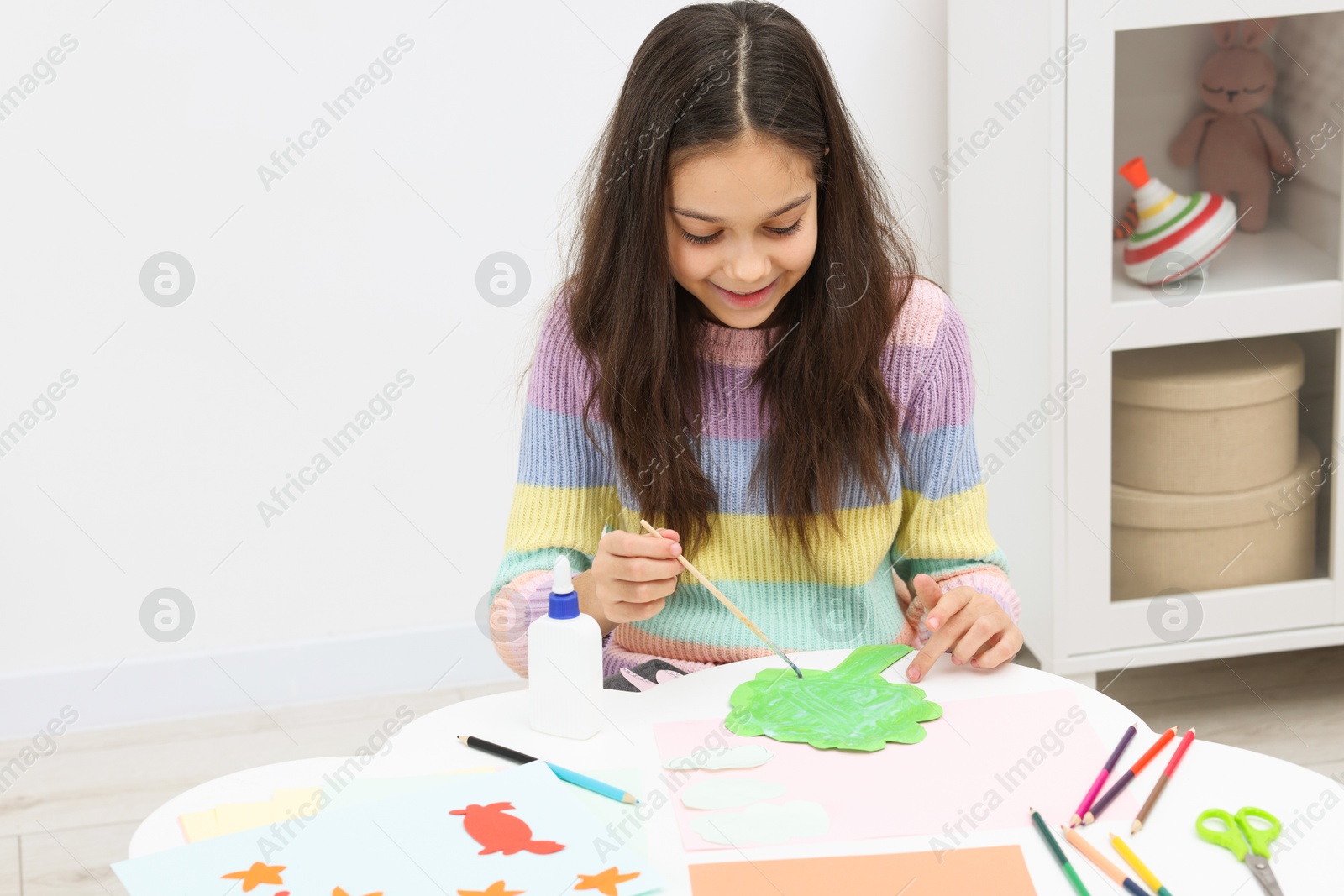 Photo of Girl applying glue onto paper figure for her creative project at table indoors. Art and craft