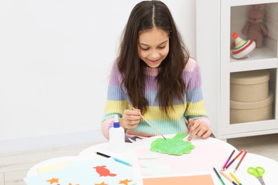 Photo of Girl applying glue onto paper figure for her creative project at table indoors. Art and craft