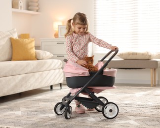 Photo of Cute little girl with her ginger cat in doll stroller at home