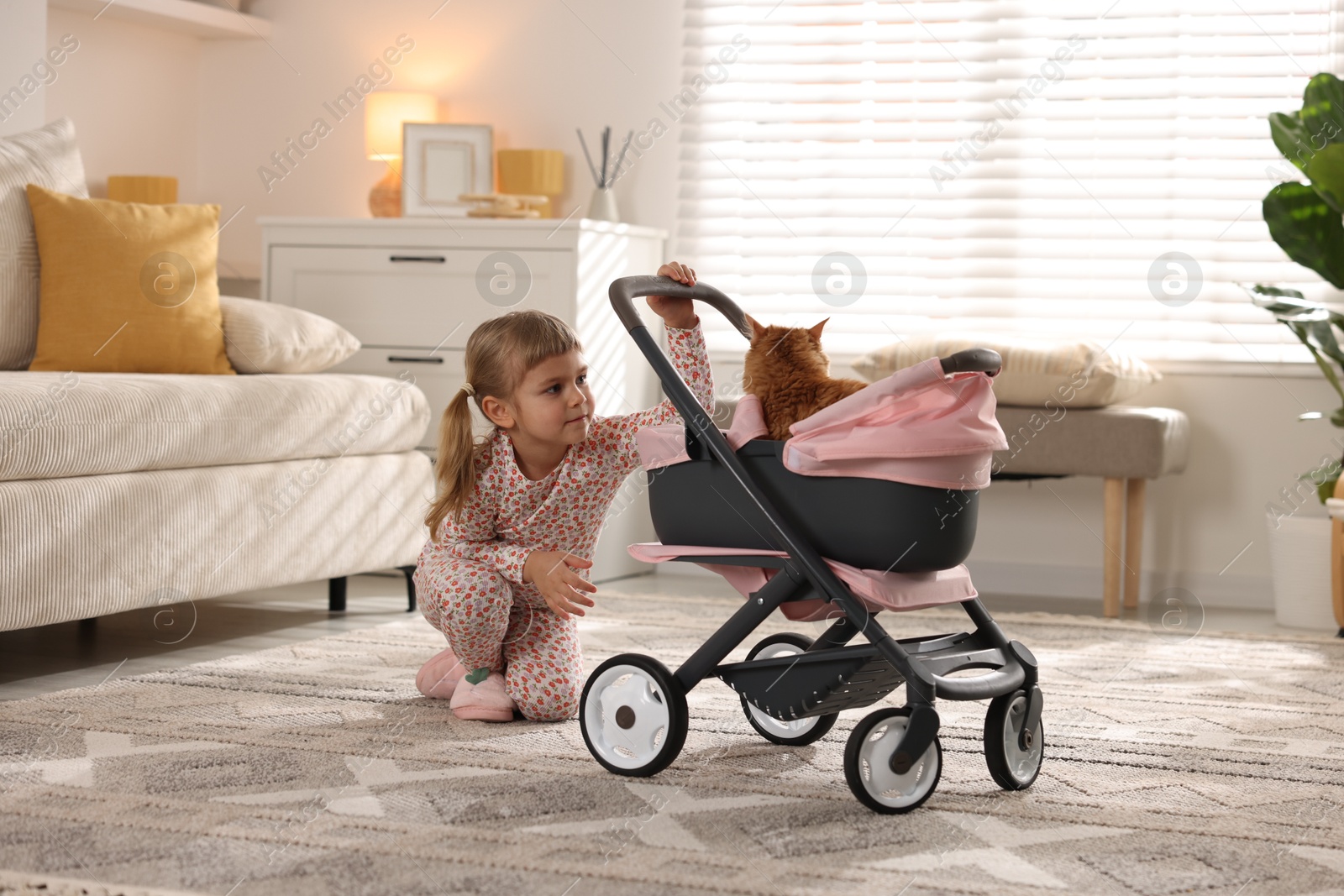 Photo of Cute little girl with her ginger cat in doll stroller at home