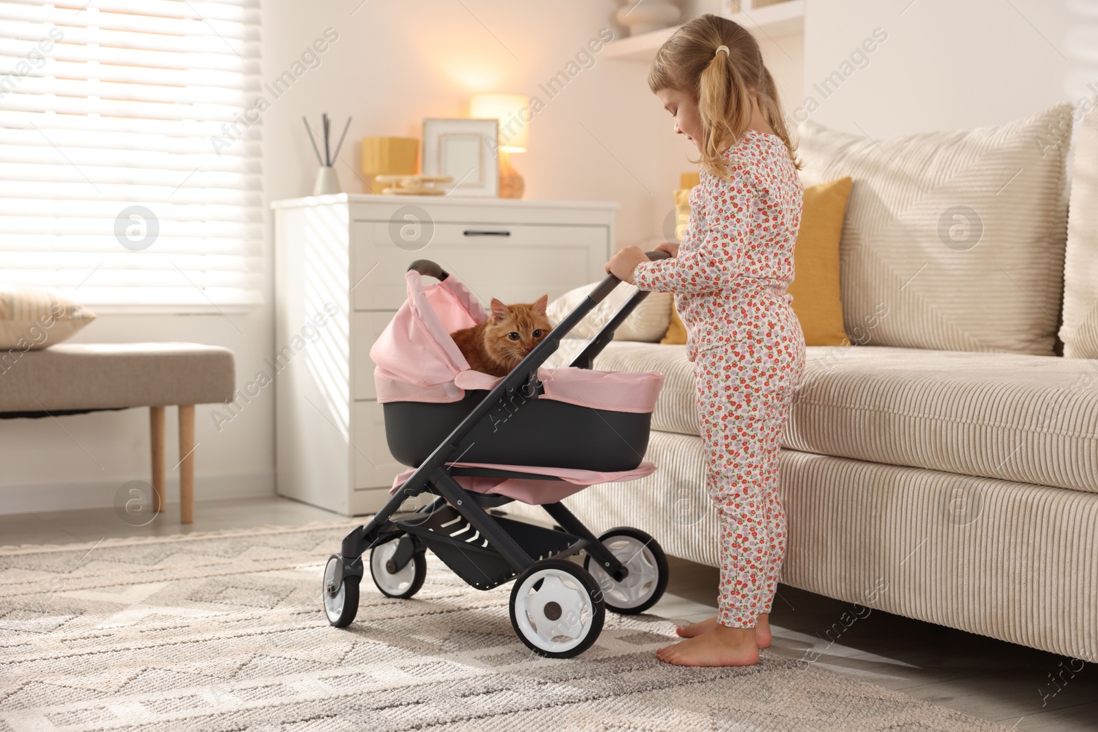 Photo of Cute little girl with her ginger cat in doll stroller at home