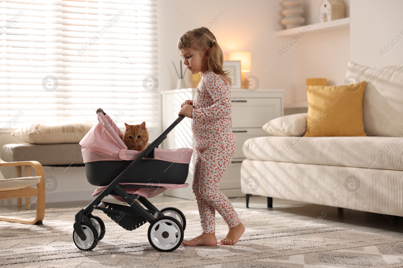 Photo of Cute little girl with her ginger cat in doll stroller at home