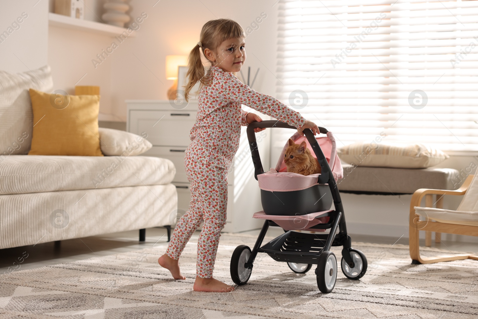 Photo of Cute little girl with her ginger cat in doll stroller at home