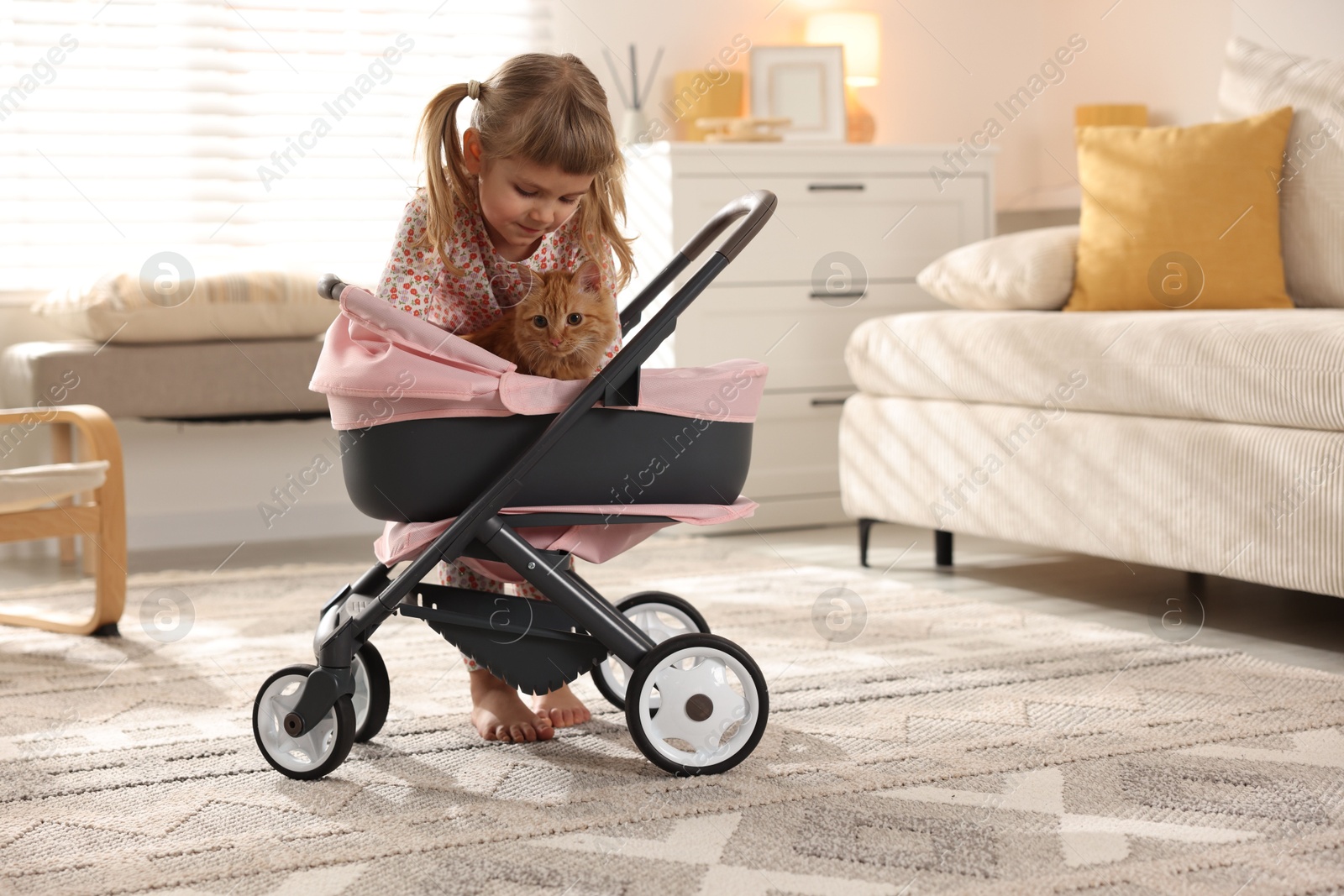 Photo of Cute little girl with her ginger cat in doll stroller at home