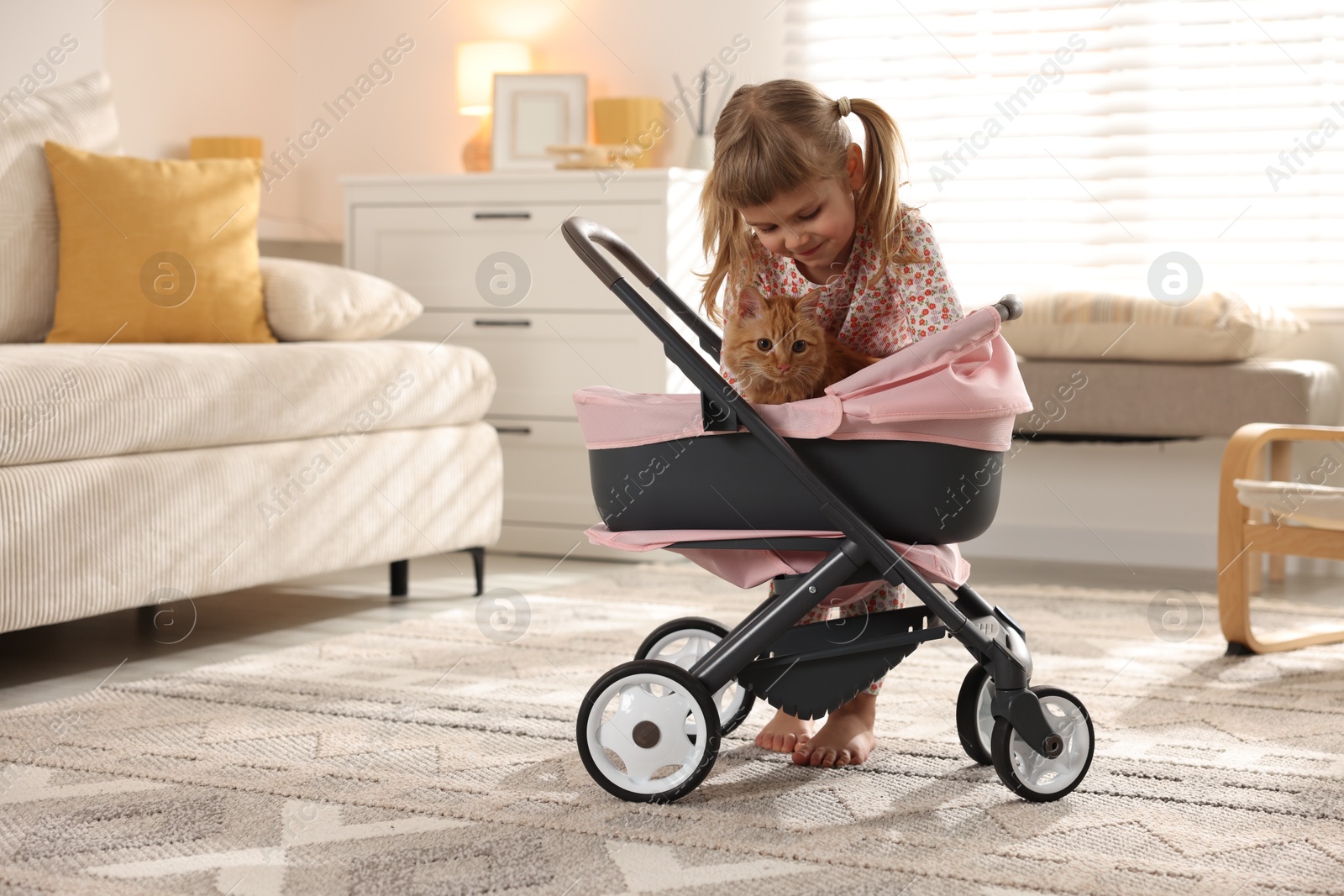 Photo of Cute little girl with her ginger cat in doll stroller at home