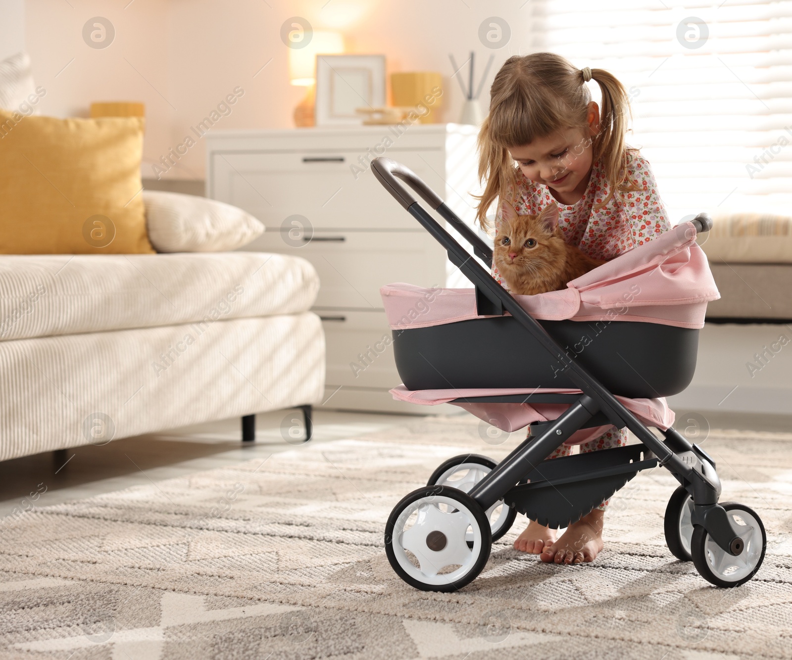 Photo of Cute little girl with her ginger cat in doll stroller at home