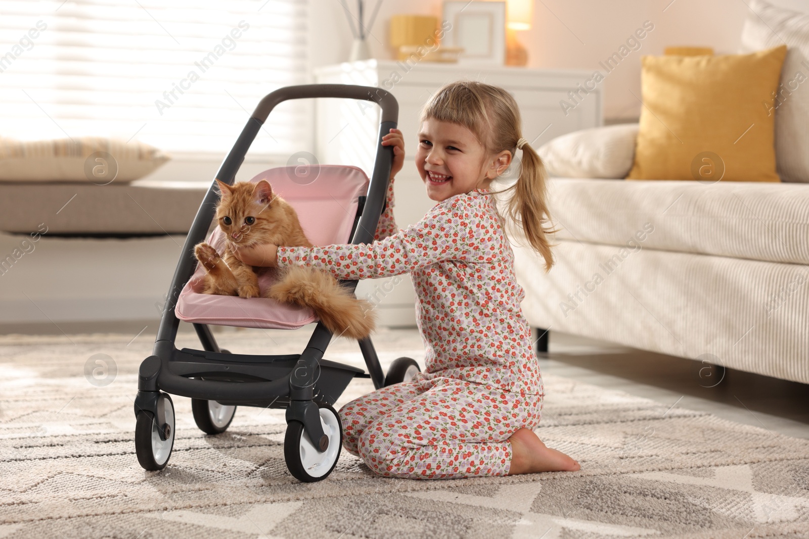 Photo of Cute little girl with her ginger cat in doll stroller at home