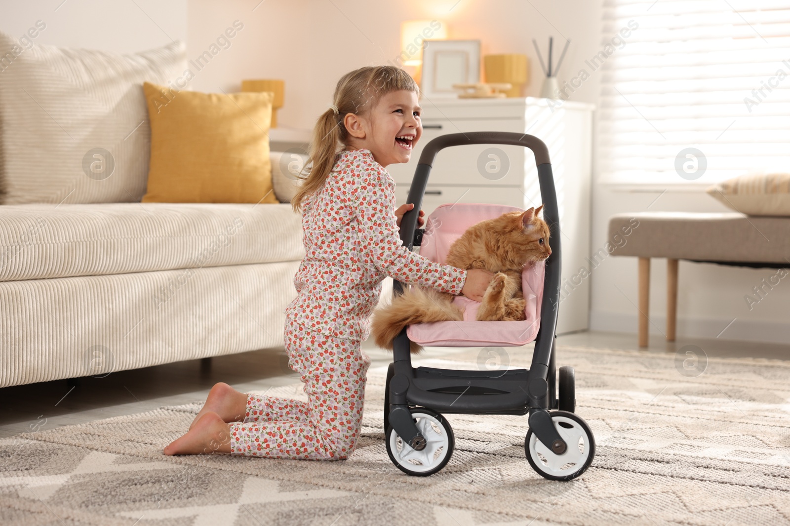 Photo of Cute little girl with her ginger cat in doll stroller at home