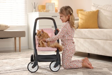 Photo of Cute little girl with her ginger cat in doll stroller at home