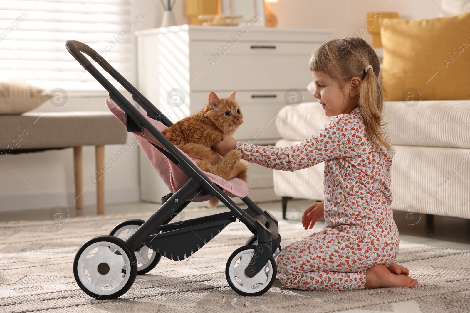 Photo of Cute little girl with her ginger cat in doll stroller at home