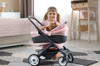 Photo of Cute little girl with her ginger cat in doll stroller at home