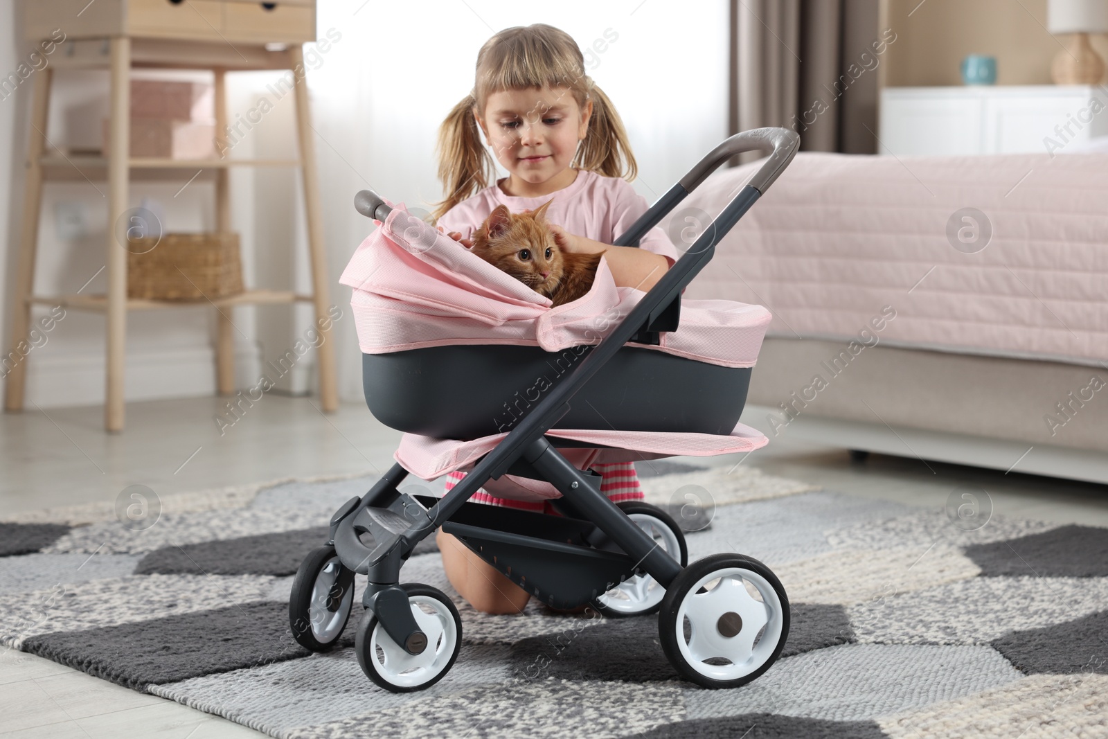 Photo of Cute little girl with her ginger cat in doll stroller at home