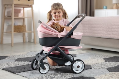 Photo of Cute little girl with her ginger cat in doll stroller at home