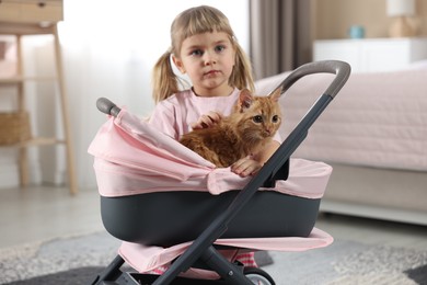 Photo of Cute little girl with her ginger cat in doll stroller at home