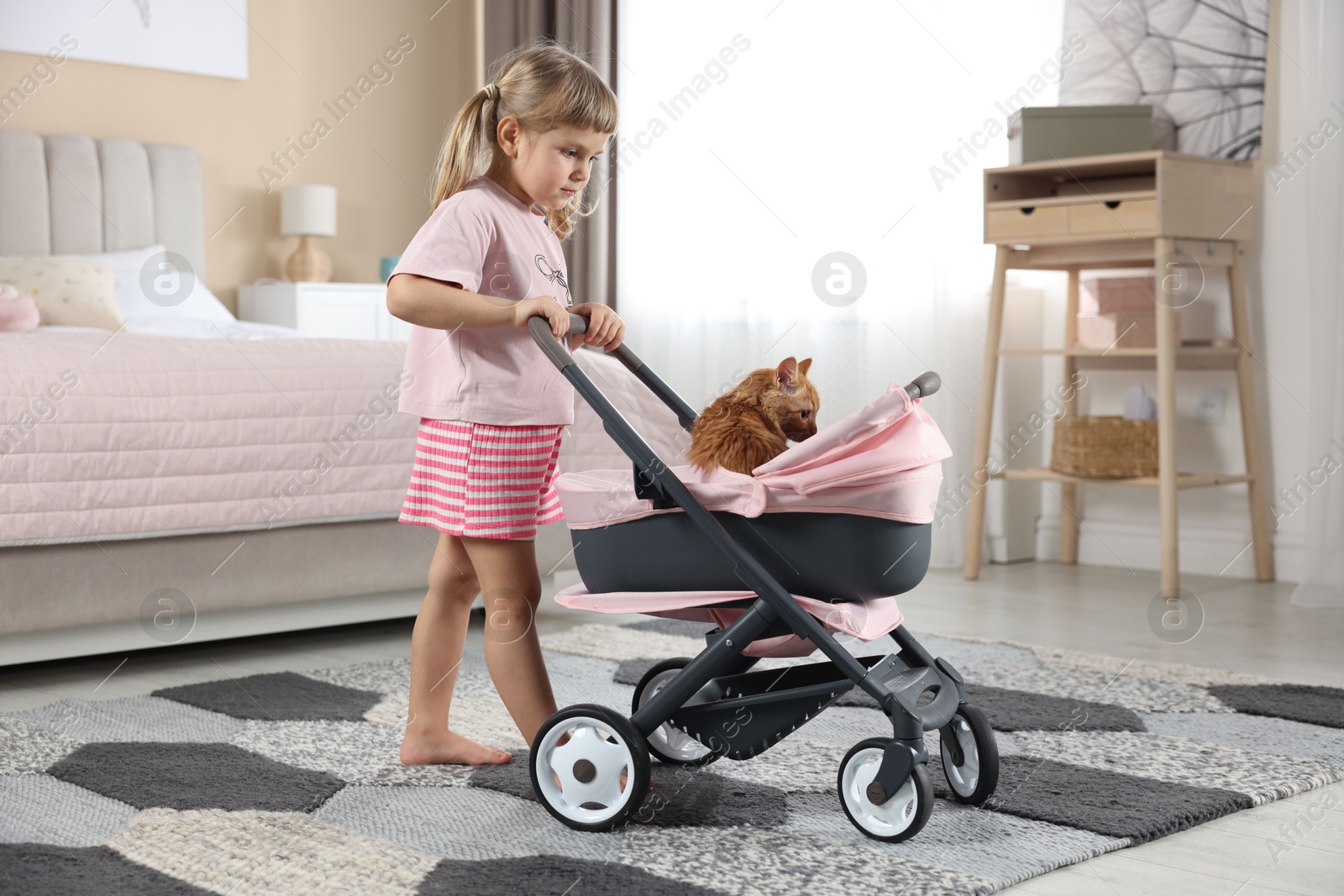 Photo of Cute little girl with her ginger cat in doll stroller at home