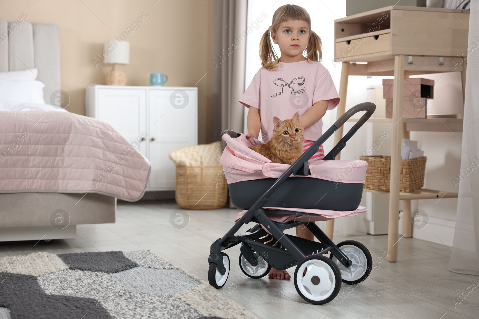 Photo of Cute little girl with her ginger cat in doll stroller at home