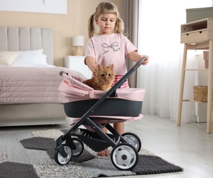 Photo of Cute little girl with her ginger cat in doll stroller at home