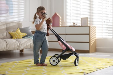 Photo of Cute little girl with doll stroller drinking from paper cup at home