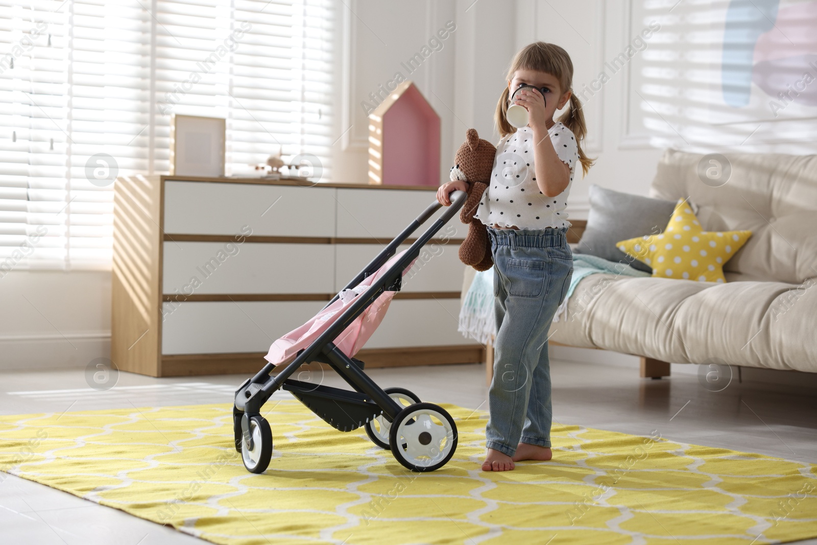 Photo of Cute little girl with doll stroller drinking from paper cup at home