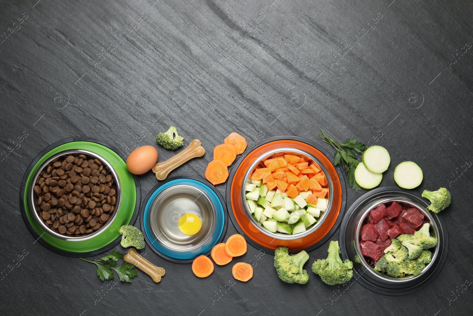 Photo of Natural and dry pet food in feeding bowls among products on dark textured table, flat lay