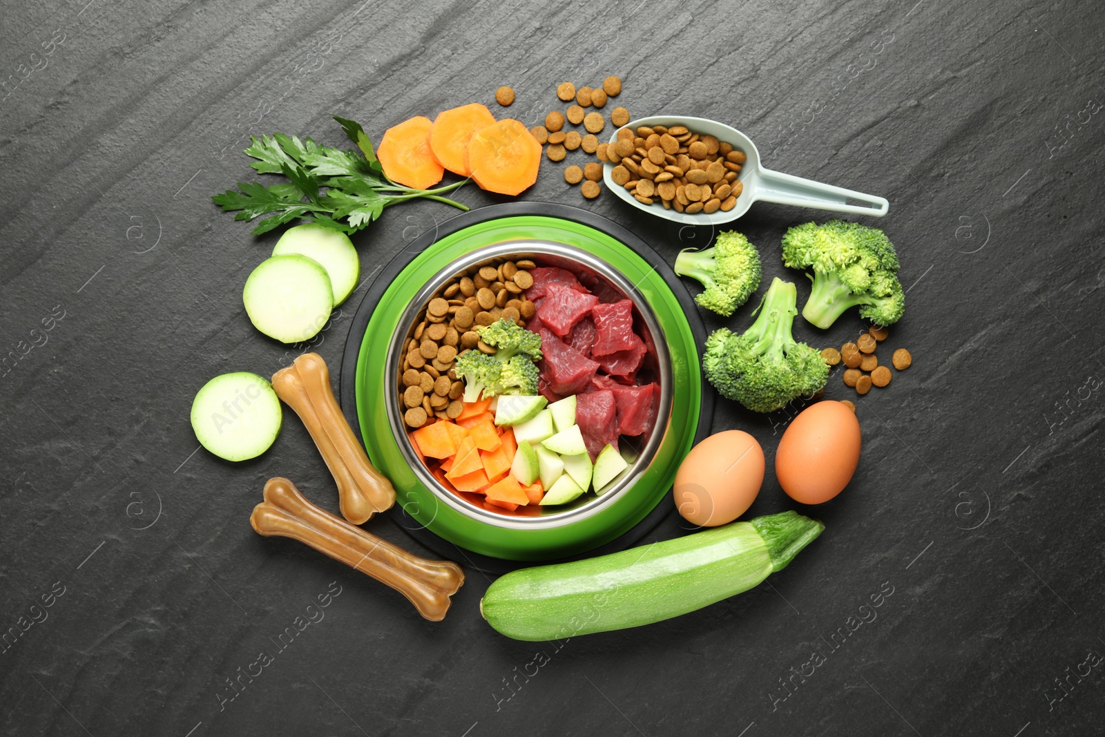 Photo of Natural and dry pet food in feeding bowl among products on dark textured table, flat lay