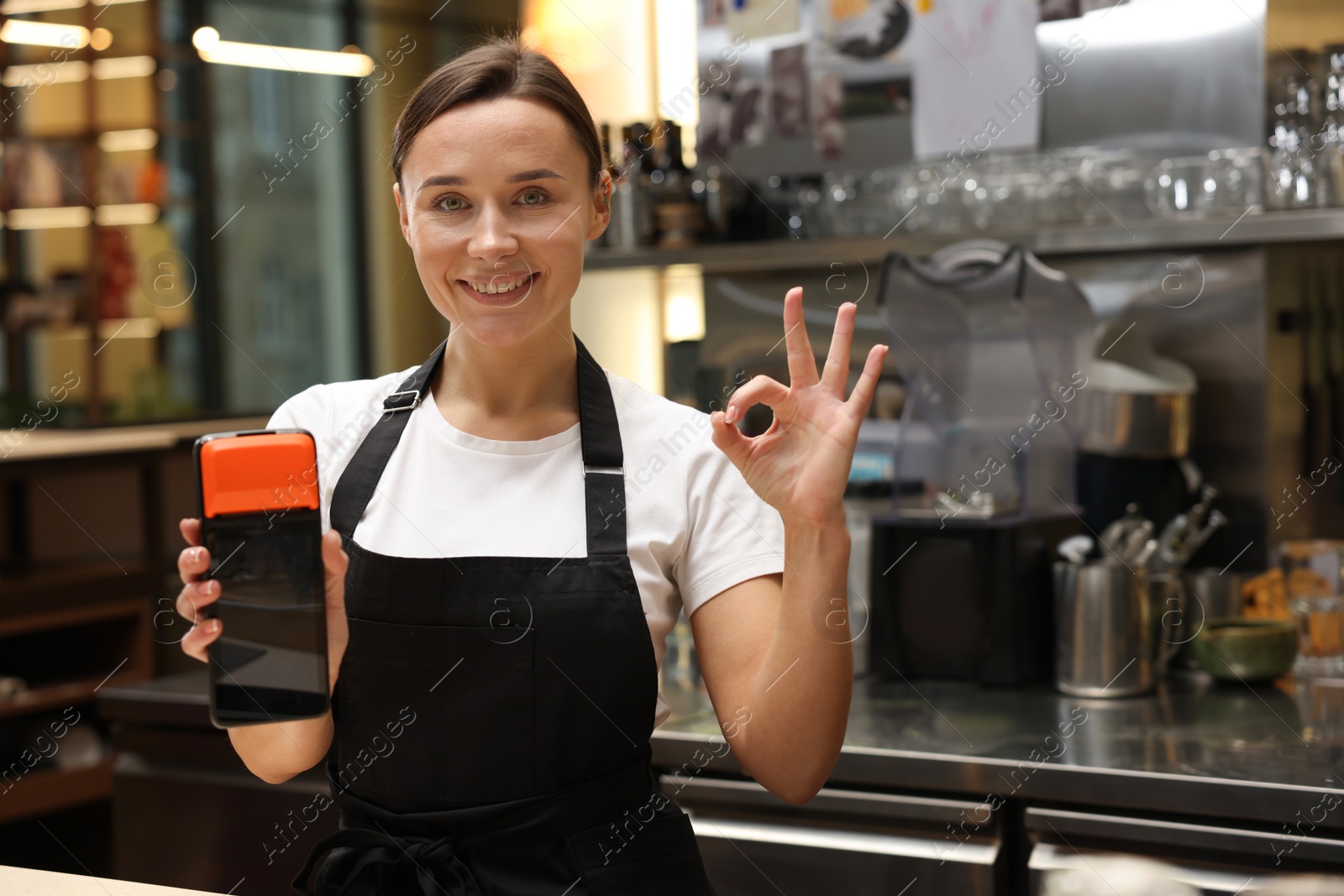 Photo of Smiling cafe worker with payment terminal showing OK gesture indoors