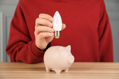 Photo of Man holding light bulb above piggy bank at wooden table, closeup. Energy saving concept