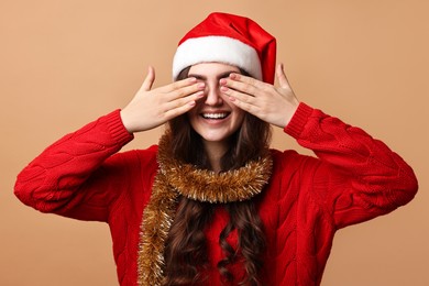 Photo of Smiling young woman with tinsel and Santa hat covering eyes on beige background