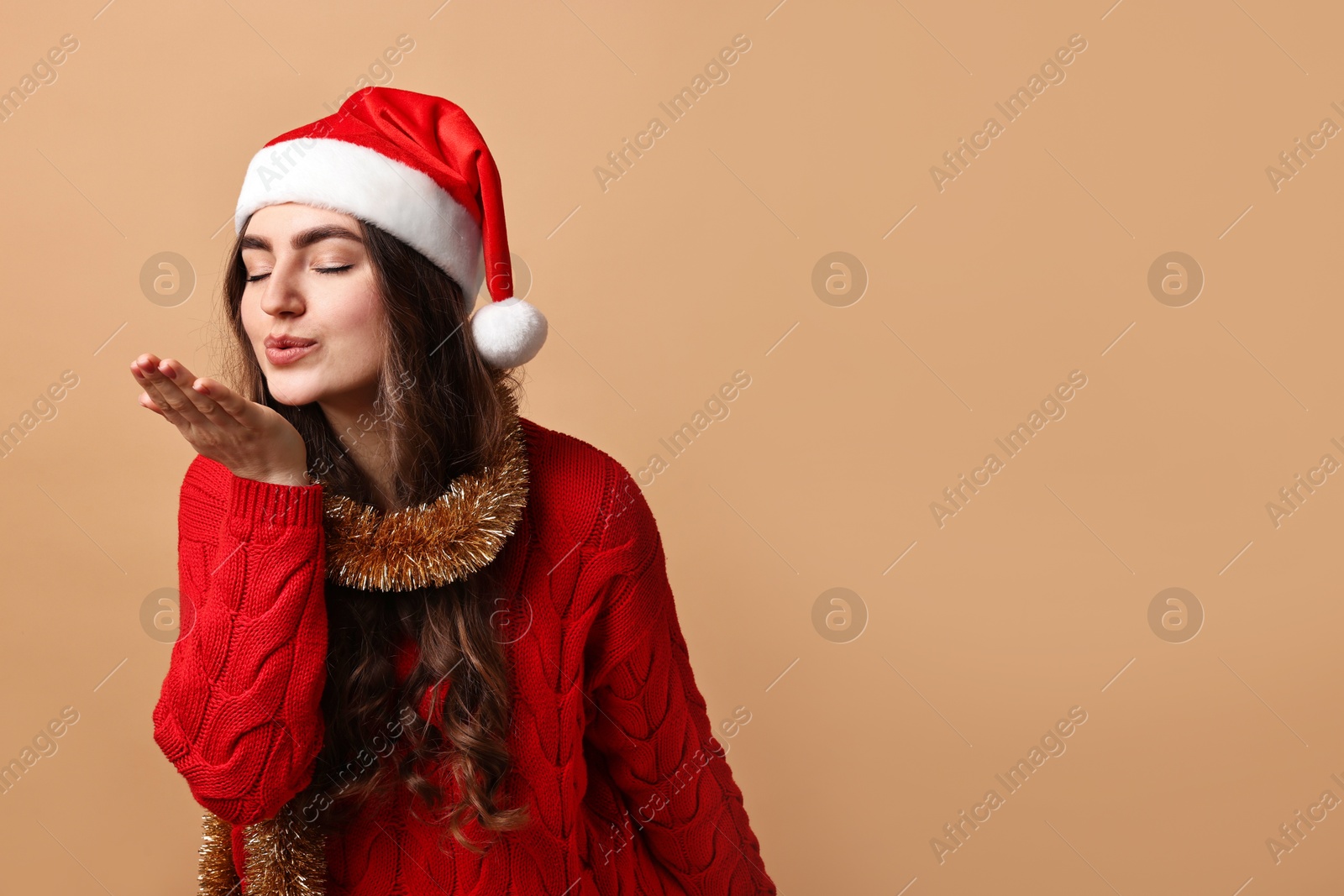 Photo of Young woman with tinsel and Santa hat blowing kiss on beige background. Space for text