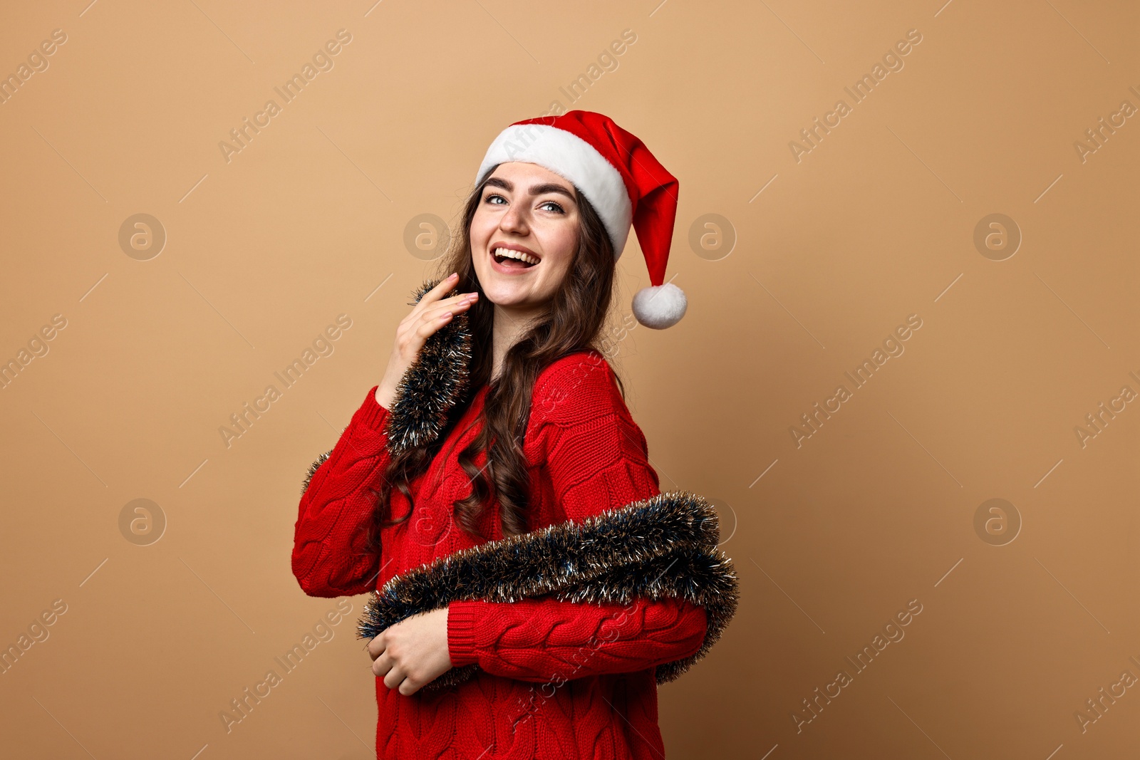 Photo of Happy young woman with tinsel and Santa hat on beige background