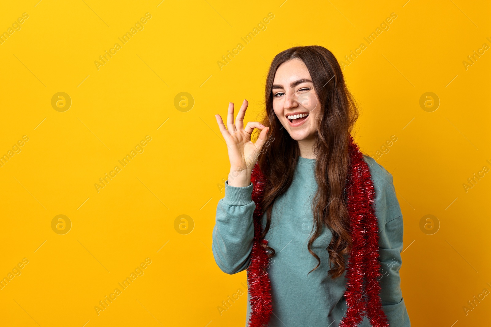 Photo of Young woman with tinsel showing OK gesture on orange background. Space for text