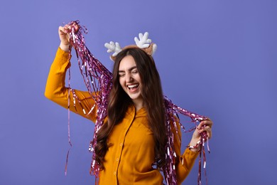 Photo of Happy young woman in reindeer headband with tinsel on purple background